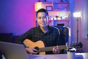 Asian man youtuber live streaming perfomance playing guitar and sing a song. Asian man teaching guitar and singing online. Musician recording music with laptop and playing acoustic guitar. photo
