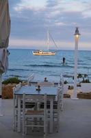 Seaside restaurant tables paddle surfer and a sailboat bathed in afternoon light in Potamos Liopetri fishing village, Mediterranean, Cyprus photo