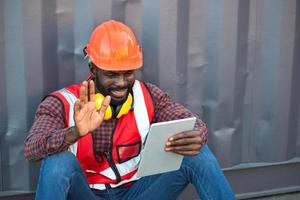 Close up happy smiling male African American engineer wearing safety vest and helmet, sitting and using mobile phone at logistic shipping cargo containers yard. photo