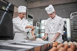 Two professional Asian male chefs in white cook uniforms and aprons are kneading pastry dough and eggs, preparing bread and fresh bakery food, baking in oven at stainless steel kitchen of restaurant. photo