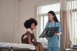 doctora asiática en controles de salud uniformes niña afroamericana, paciente con enfermedad, diagnóstico explica la película de rayos X en la cama de la sala de emergencias en la sala del hospital, clínica pediátrica, consulta de examen de niños. foto