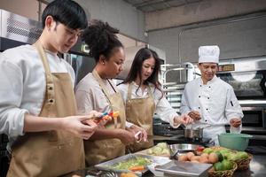 Hobby cuisine course, senior male chef in cook uniform teaches young cooking class students to peel and chop apples, ingredients for pastry foods, fruit pies in restaurant stainless steel kitchen. photo