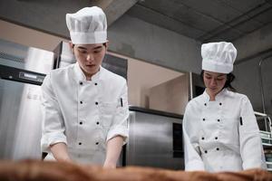 Two professional Asian chefs in white cook uniforms and aprons knead pastry dough and eggs, prepare bread, cookies, and fresh bakery food, baking in an oven in a restaurant stainless steel kitchen. photo