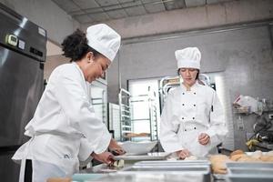 dos chefs profesionales con uniformes y delantales de cocina blanca amasan masa de pastelería y huevos, preparan pan, galletas y comida fresca de panadería, horneando en horno en una cocina de acero inoxidable de un restaurante. foto