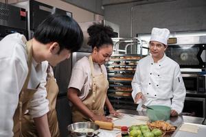 Cuisine course, senior male chef in cook uniform teaches young cooking class students to knead and roll pastry dough, prepare ingredients for bakery foods, fruit pies in stainless steel kitchen. photo