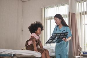 doctora asiática en controles de salud uniformes niña afroamericana, paciente con enfermedad, diagnóstico explica la película de rayos X en la cama de la sala de emergencias en la sala del hospital, clínica pediátrica, consulta de examen de niños. foto