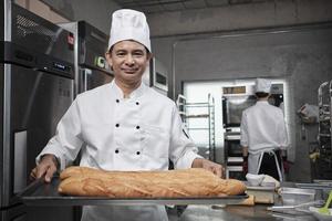 Senior Asian male chef in white cook uniform and hat showing tray of fresh tasty bread with a smile, looking at camera, happy with his baked food products, professional job at stainless steel kitchen.