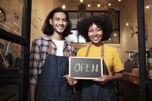 Two young startup barista partners with aprons stand at casual cafe door, letters on board and show open sign, happy and cheerful smiles with coffee shop service jobs, and new business entrepreneurs. photo