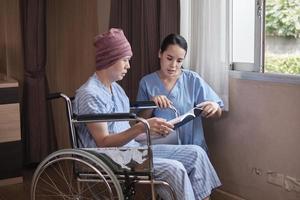 Uniformed young Asian female therapy doctor motivates wheelchair male patient at window by reading book to support recovery cancer illness after chemo medical treatment in hospital inpatient room. photo