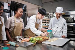 Cuisine course, senior male chef in uniform teaches young cooking class students, brushes pastry dough with eggs cream, prepares ingredients for bakery foods, fruit pies in stainless steel kitchen. photo