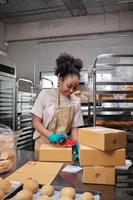 entrega de pequeñas empresas de inicio de panadería. una cocinera afroamericana está empacando pan y pasteles hechos a mano y recién horneados en cajas y enviando para compras de clientes en línea en la cocina culinaria. foto