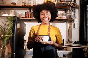 African American female barista in looks at camera, offers cup of coffee to customer with cheerful smile, happy service works in casual restaurant cafe, young small business startup entrepreneur. photo