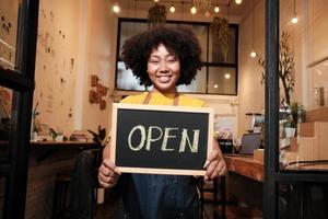 One African American female startup barista stands at casual cafe door, looks at camera, and shows open sign, happy and cheerful smiles with coffee shop service jobs, and new business entrepreneurs. photo