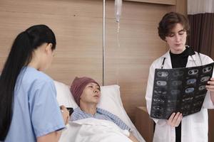 Caucasian female doctor in uniform diagnosis explains x-ray film with Asian radiologist and recovery male patient at inpatient room bed in a hospital ward, medical clinic, cancer examination consult.