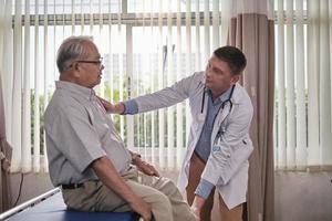 Caucasian male doctor in uniform health checks illness older patient, physical therapy on legs at emergency room bed in a hospital ward, orthopedics and medical clinic, body pain examination consult. photo