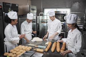Multiracial professional gourmet team, four chefs in white cook uniforms and aprons knead pastry dough and flour, prepare bread, and bakery food, baking in oven at stainless steel restaurant kitchen. photo