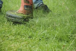 Close-up shot of male worker in jeans maintenance garden using an electric lawnmower to trim overgrown green grass in the lawn yard, scattering the grass in summer, outdoor landscaping work service. photo