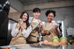 tres jóvenes estudiantes en clase de cocina usan delantales disfrutan y son alegres y divertidos con ingredientes y herramientas en la cocina, sonríen felices, preparan frutas para aprender juntos un curso culinario divertido. foto