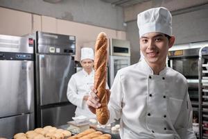 Portrait of young Asian male chef in white cooking uniform looks camera, happy smile and cheerful with baguette, professional foods occupation, commercial pastry culinary jobs in a restaurant kitchen. photo