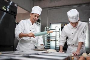 Two professional Asian male chefs in white cook uniforms and aprons are kneading pastry dough and eggs, preparing bread and fresh bakery food, baking in oven at stainless steel kitchen of restaurant. photo