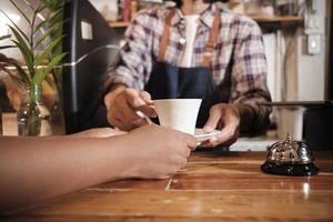 Front close-up of barista's hand works by serving white cup of hot coffee to customer order at wooden counter bar in casual cafe in morning, modern lifestyle works service job in coffee shop business. photo