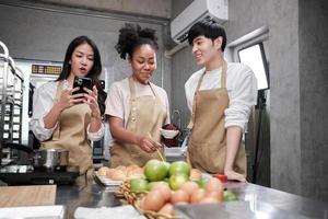 tres jóvenes estudiantes en la clase de cocina usan delantales para disfrutar mientras toman una foto selfie con el teléfono móvil en la cocina, sonríen y ríen, preparan huevos y frutas, aprenden juntos un curso culinario divertido.