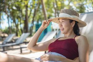 Model Asian woman wearing red swimsuit and straw hat lying on the sun lounger in sun by the sea. Beautiful girl relaxing at infinity luxury resort. Spa, wellness photo