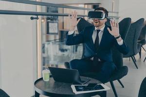 Businessman dressed in formal suit wearing VR goggles while working on laptop in cafe photo