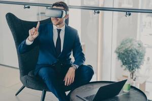 Smiling businessman using VR headset glasses while sitting in office lobby and working on laptop photo