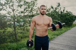 hombre musculoso guapo levanta pesas afuera, hace entrenamiento de bíceps, se para con el torso sexy desnudo, entrena los músculos, vestido con pantalones cortos posa al aire libre, tiene un cuerpo atractivo. concepto de levantamiento de pesas foto