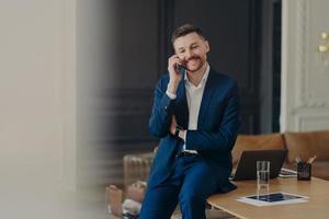 Happy handsome businessman talking on phone while sitting on office desk photo