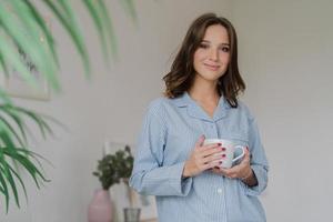 Waist up shot of pleasant looking young woman dressed in striped pyjamas, drinks hot beverage after awakening, has sparre time at home, stands indoor against cozy interior. People and rest concept photo