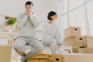 Worried couple pose near stack of cardboard boxes, have to move in other flat, have no money for rent, have dejected sad expressions, sit together in white room, have family conflict and divorce photo
