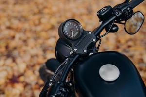 Close up shot of black motorbike against ground covered with orange foliage in autumn park. Transportation, riding and traveling concept. photo