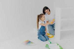 Little girl learns how to paint with roller, stands on knees, her mother tells how to do it correctly, pose in spacious white room, choose color from palette samples. Home makeover, decoration concept photo