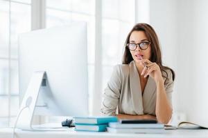 Contemplative female entrepreneur keeps pen in mouth, focused in monitor of computer, thinks on development of new strategy, wears elegant clothes and spectacles, poses against office interior photo