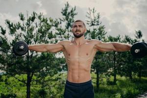 Strong healthy man stretches arms, makes weightlifting and exercises biceps with barbells, poses with naked muscular torso outdoor. Athletic shirtless motivated sportsman has workout in open air photo