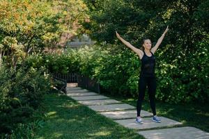 una foto completa de una deportista activa levanta los brazos respira profundamente vestida con ropa activa tiene entrenamiento deportivo en el parque durante la mañana escucha música en los auriculares. concepto de estilo de vida deportivo.