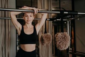 mujer joven en forma en ropa deportiva negra tomando un descanso en la máquina de pilates entre entrenamientos en el gimnasio foto