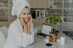 Pretty smiling young female model applies collagen patches under eyes to reduce wrinkles wears bathrobe and towel on head enjoys skin care procedures sits at desk with cosmetic tools cozy interior photo