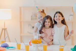 Horizontal shot of three happy friends embrace and have funny expressions, pose near festive table with cake indoor. Three girls celebrate birthday together, have fun, come to party photo