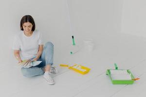 Satisfied brunette woman chooses color from samples, going to refurbish room, sits on floor in casual clothes, surrounded with trays and paint rollers, busy with house renovation. DIY concept photo