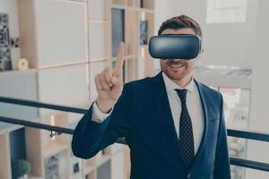 Young cheerful businessman in formal suit and VR headset goggles standing in company lobby photo