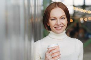 Good looking young female dressed in casual white turtleneck sweater, holds paper cup of aromatic cappucino or coffee, looks happily at camera, poses outside, has spare time. Lifestyle concept photo