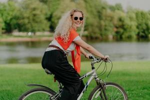 Photo of attractive young woman with curly hair, rides bicycle, wears sunglasses, t shirt, pants, jumper on shoulders, enjoys summer time, fresh air outdoor, spends free time actively with friends