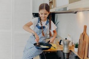 Little girl with pigtails wears apron, learns to cook, poses near stove, prepares fried eggs for breakfast, helps parents with cooking, busy at modern kitchen. Children, culinary, food concept photo