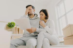 Dejected husband and wife manage finances, receive bill, face financial problem, have gloomy expressions, sit together in empty room, big window behind, cardboard boxes with personal stuff near photo