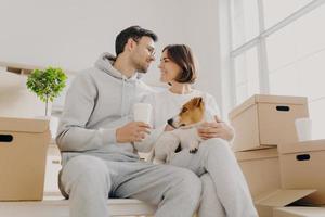 Indoor shot of affectionate woman and man express love to each other, have good relationship, drink coffee, pose with favourite pet, have to unpack many cardboard boxes, bought new apartment photo