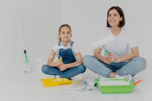 Photo of cheerful girl and daughter sit crossed legs on floor, hold paint rollers, paint room in white color, busy with refurbishment, have happy expressions. Home, repair, building and decoration