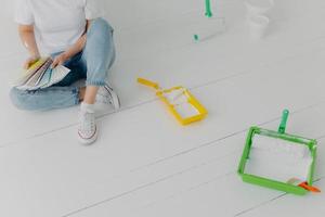 Cropped image of faceless woman chooses color from samples, going to refurbish walls in own room, wears sneakers, sits on white wall with trays and paint rollers. Repair and designing concept photo